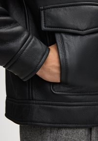 Zwarte leren jas met een textuurafwerking, voorzien van een grote patchzak en gestikte details op de mouwen en onderkant.