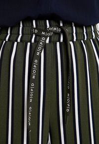 Gröna och vita randiga byxor med dragsko i midjan som har ett svart band med vit text. Mellantungt tyg, avslappnad passform.