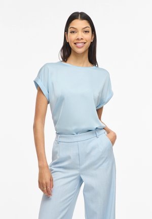 Mladá žena sa usmieva, má na sebe svetlomodrú blúzku s krátkym rukávom a zodpovedajúce vysoké nohavice, stojí s jednou rukou v vrecku.