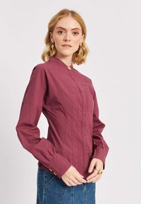 Donna con capelli biondi che indossa una blouse attillata a maniche lunghe color marrone scuro con bottoni dorati e orecchini pendenti dorati, in piedi davanti a uno sfondo semplice.