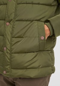 Olivengrøn polstret jakke med en glat tekstur, der har knaplukning og sidelommer. Syningerne er synlige på stoffet.