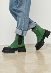 Groene suede enkellaarsjes met zwarte accenten, stevige rubberen zool, een zijrits en elastische inzetten. Gedragen met opgerolde lichtblauwe spijkerbroeken.