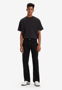Zwarte katoenen t-shirt met korte mouwen en reliëflogo, gecombineerd met zwarte jeans en witte schoenen met zwarte accenten.
