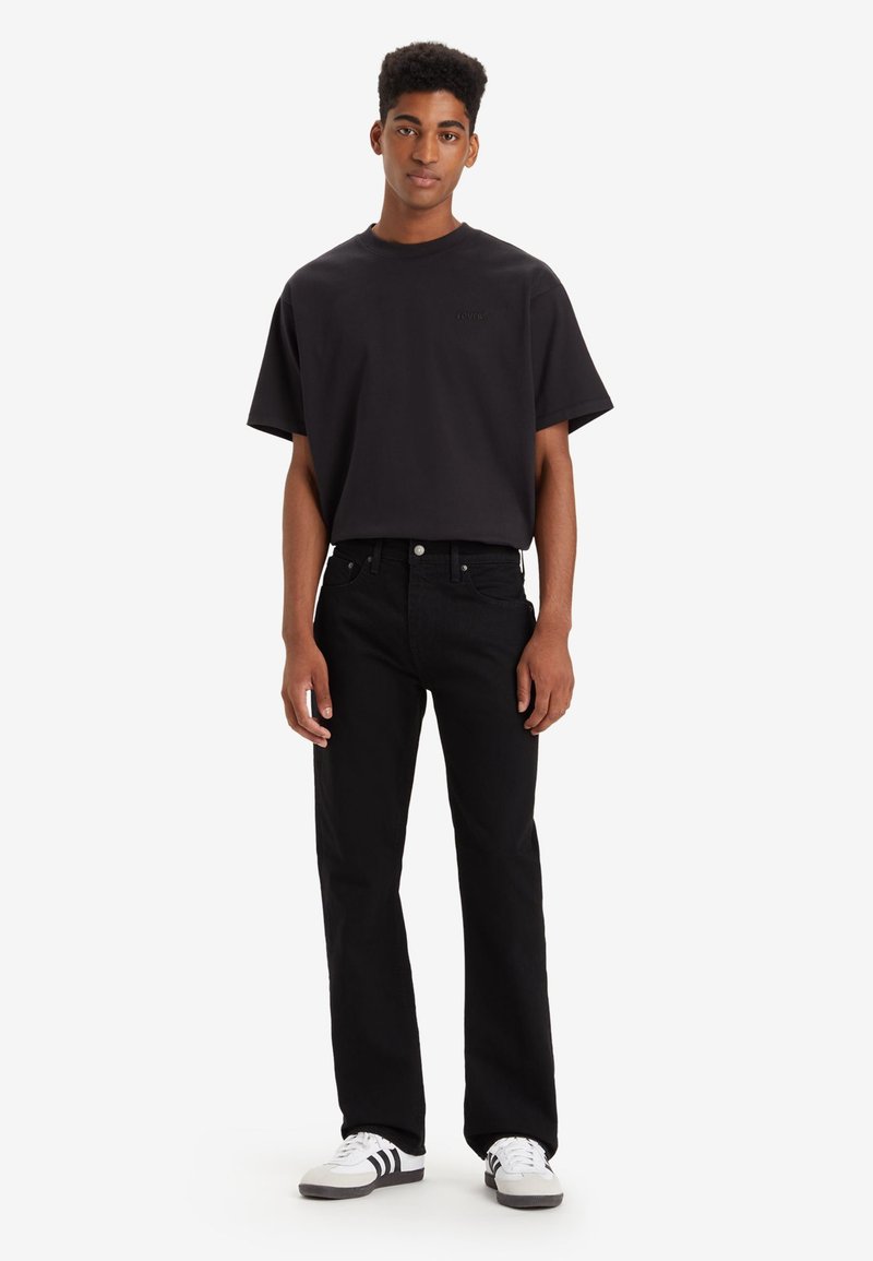 Schwarzes Baumwoll-T-Shirt mit kurzen Ärmeln und eingeprägtem Logo, kombiniert mit schwarzen Jeans und weißen Schuhen mit schwarzen Akzenten.