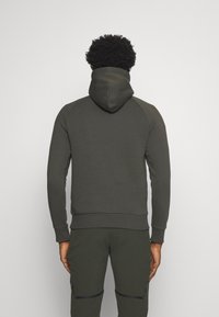 Uomo di spalle che indossa un cappuccio oliva scuro e pantaloni coordinati con tasche con zip, in piedi contro uno sfondo grigio chiaro e semplice.