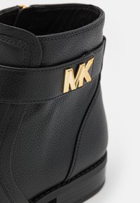 Gros plan d’une botte en cuir noir texturé avec un logo MK doré sur la bride de cheville et un petit talon empilé.