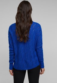 Blauer Strickpullover mit einem strukturierten Spitzenmuster, mit langen Ärmeln und lockerem Schnitt, kombiniert mit schwarzer Hose.