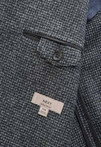Grå vævet blazer med en struktureret stof, sort kant omkring lommen, og et mærke der står "NEXT REGULAR FIT" med størrelsesoplysninger.