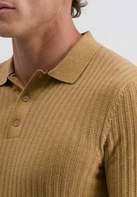 Strick-Polohemd in einer warmen Sandfarbe mit strukturiertem Rippmuster. Verfügt über einen klassischen Kragen und eine Knopfleiste mit zwei Knöpfen sowie dezenten Akzenten.