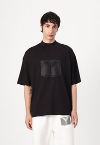 Zwarte T-shirt met een hoge hals, oversized pasvorm, voorzien van een vierkante graphic met strepen en de tekst "CARLO COLUCCI" in het midden.