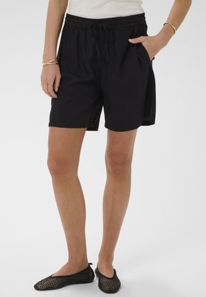 Person iført sorte shorts med snøre og sorte slip-on sko i mesh, med den ene hånd i lommen og gyldne armbånd om håndleddene.