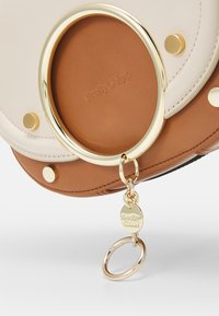 Borsa in pelle marrone chiaro e crema con un accento in metallo dorato circolare, dotata di un portachiavi con un'etichetta del logo e borchie dorate piatte.