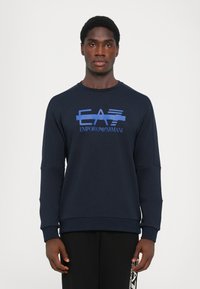 Navy zila džemperis, kas izgatavots no mīksta auduma, ar zilu logo dizainu "EA7 EMPORIO ARMANI" uz priekšpuses un garām piedurknēm.