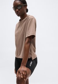 Femme portant des lunettes de soleil, un t-shirt marron ample et un short de sport noir avec le logo "TERREX", posant avec une main sur le genou devant un fond blanc.