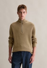 Beiger Strickpullover mit hohem Reißverschlusskragen, gerippter Textur und langen Ärmeln. Stilisiert mit dunklen Jeans. Einfaches, funktionales Design.