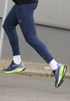 Leggings atléticos en tono azul marino con acentos reflectantes y un zipper lateral. Un par de zapatillas de correr en gris y verde neón con parte superior texturizada y suela acolchada.