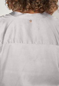 Blouse en satin gris clair avec un col arrondi, un détail froncé dans le dos et un petit logo métallique au niveau du cou. Texture lisse.