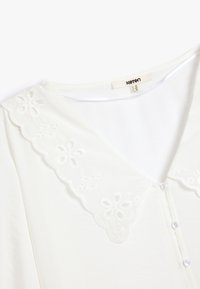 Blusa bianca con scollo a V e colletto in pizzo ricamato. Presenta una chiusura a bottoni con piccoli bottoni traslucidi e una trama di tessuto morbido.