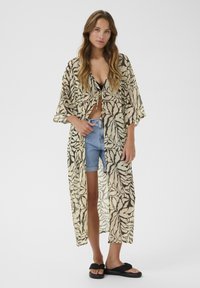 Kimono long et transparent avec des motifs de feuilles beiges et noires, doté de larges manches et d'une ceinture à la taille, porté sur un short et des sandales.