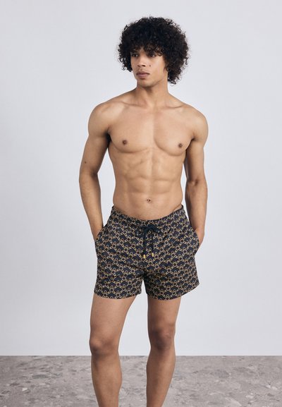 Muskuļots vīrietis ar cirtainiem matiem, ģērbies melnās peldbiksēs ar brūnu ķēdes rakstu, stāv ar rokām kabatās uz akmens grīdas.