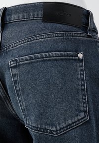 Jean en denim bleu foncé présentant une poche arrière avec un bouton en métal, une étiquette en cuir noir au niveau de la taille, et des détails de couture visibles.