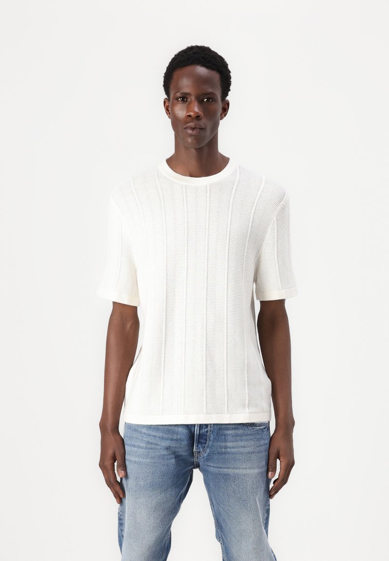 Weißes, geripptes Strick-T-Shirt mit kurzen Ärmeln und rundem Halsausschnitt, das horizontale und vertikale Muster aufweist, kombiniert mit hellblauen Jeans.