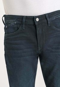 Jeans bleu foncé en denim, avec une coupe droite, un design à cinq poches et une fermeture à bouton et zip à l'avant. Texture légèrement décolorée.