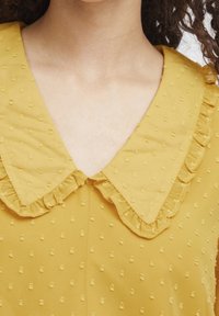 Senfgelbes Kleid mit texturierter Oberfläche, ausgestattet mit einem Rüschenausschnitt und einem subtilen Punktmuster über den gesamten Stoff.