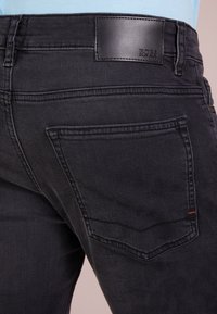 Detailní záběr na zadní kapsu černých džínů z denimu s koženou nášivkou značky BOSS na pase, nošených s světlomodrou košilí.