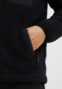 Sudadera de fleece azul marino con una superficie texturizada, un bolsillo en el pecho negro y un dobladillo elástico. Incluye bolsillos laterales con cremalleras.