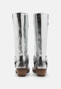 Bottes en cuir argenté montantes avec une finition texturée, dotées d'un talon en bois brun et de sangles métalliques avec des clous. Vue de dos.