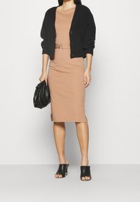 Cardigan noir court en maille porté sur une robe beige ajustée, longueur genou, avec une taille ceinturée, accompagné de mules noires à bout ouvert et d'une pochette noire.