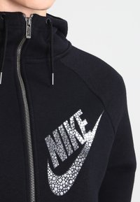 Svart zip-up hoodie med en strukturerad silverfärgad Nike-logotyp med rhinestone-effekt på bröstet, kontrasterande svart tyg och huva med dragsko.