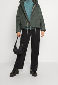 Grön puffjacka med huva, lager-på-lager över en teal stickad tröja, i kombination med svarta vida byxor och chunky svarta stövlar. Håller en svart väska.