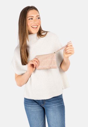 Kvinne iført hvit genser og blå jeans, smilende mens hun holder en liten beige, quiltet håndveske med glidelås.