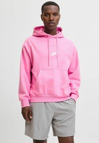 Sudadera rosa de tela suave con un bolsillo frontal, cordón ajustable y un pequeño logotipo blanco en el pecho. Combinada con pantalones cortos grises.
