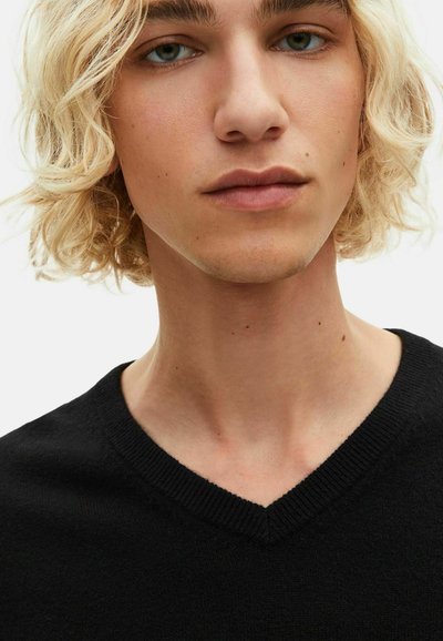 Schwarzer V-Ausschnitt-Pullover aus weichem Strickstoff. Nahaufnahme des Gesichts einer jungen Person mit leicht welligem Haar und neutralem Make-up, das eine klare Haut zeigt.