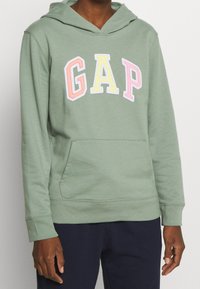 Sudadera verde con capucha, bolsillo frontal y logo "GAP" bordado en colores pastel. Material de algodón suave con un ajuste cómodo y capucha.