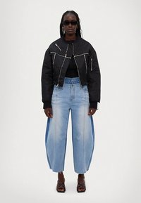 Kvinde med fletninger iført sort cropped jakke med sølv lynlåse, vidde tofarvede blå jeans, sorte strop sandaler og solbriller.