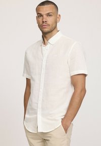 Chemise en lin blanche à manches courtes avec un col boutonné et une texture subtile. Présente un petit logo sur la poitrine gauche et un ourlet droit.