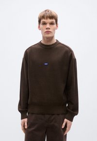 Pull en maille marron avec encolure et ourlet côtelés. Présente un petit logo rectangulaire bleu sur la poitrine. Manches longues et coupe décontractée.