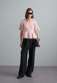 Světle růžový, huňatý top s krátkým rukávem spárovaný s černými, volnými kalhotami. Modelka drží černou kabelku, nosí černé sluneční brýle a minimální doplňky.