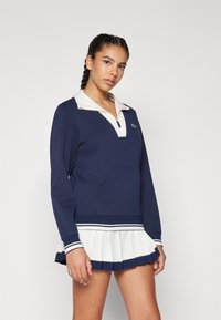 Marinblå tröja med vit dragkedja vid kragen och ribbade muddar, utrustad med en framficka och en vit plisserad kjol med marinblå ränder.