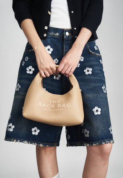 Des mains tenant un sac à main en cuir beige étiqueté "THE SACK BAG MARC JACOBS" au-dessus de shorts en denim avec une broderie de sequins floraux blancs.