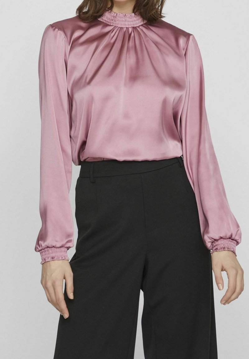 Rosa satinblus med hög hals och rynkdetalj, långärmade med elastiska manschetter, i kombination med svarta byxor. Slät textur.