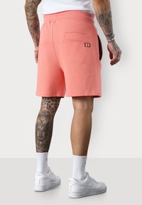 Shorts en sweat rose corail avec une taille élastique, des poches latérales et une poche arrière portant un logo noir. Associés à des baskets et des chaussettes blanches.