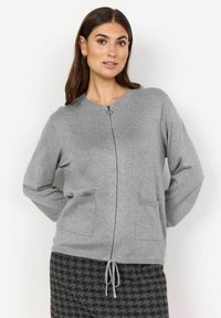 Grå zip-up kofta i mjukt material, med långa ärmar, sidofickor och rund halsringning. Dragsko detalj vid fållen.