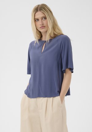 Kobieta z długimi blond włosami, ubrana w luźną, niebieską bluzkę z krótkim rękawem i beżowe spodnie, z rękami w kieszeniach, patrząca w bok.