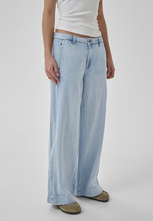 Osoba ubrana w jasnoniebieskie dzwony, beżowe buty i białą koszulkę bez rękawów na jednolitym tle.