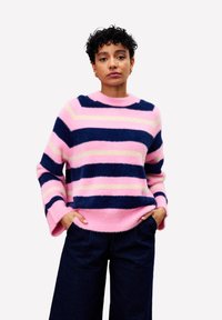 Jeune personne avec des cheveux courts et bouclés portant un pull en laine rayé rose et marine et un pantalon large noir, les mains dans les poches, expression neutre.